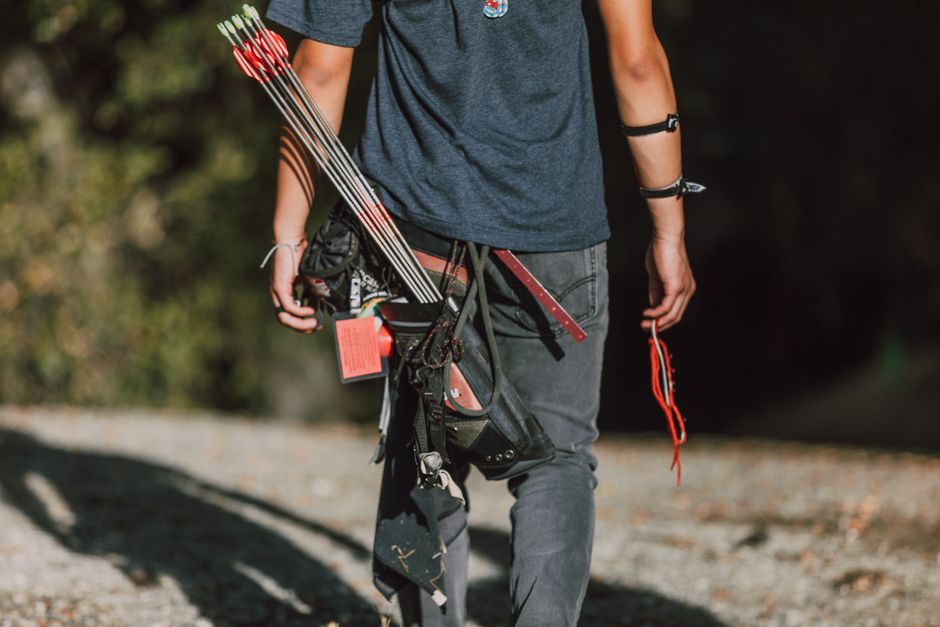 A person dressed in a dark grey t-shirt and grey trousers is seen from the back, walking outdoors on a gravel surface during daylight. They are carrying a black tool belt around their waist filled wit