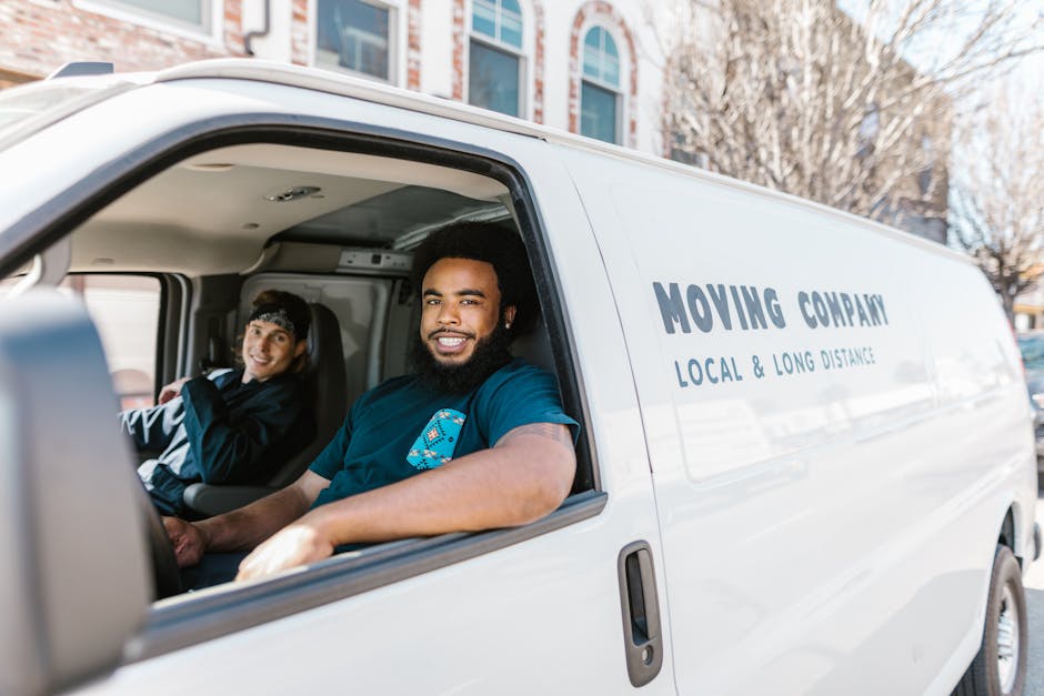 Inside a white moving van from Man and Van Bow, two movers are visible preparing for a home relocation. The driver, a man with a beard and short dark hair, is smiling at the camera, wearing a blue uniform with a logo on the chest, seated in the driver’s seat. Next to him, a woman with long dark hair, wearing a dark zip-up jacket, is seated in the passenger seat, also smiling. The van’s interior shows safety equipment such as seat belts, and the vehicle exterior displays the company's name, 'Moving Company,' along with the services offered, including local and long-distance moves. The scene is set outdoors with a background of urban buildings, and sunlight illuminates the scene, emphasizing the professional moving process involving packing supplies, such as cardboard boxes and protective blankets, inside the van, which is part of the furniture transport and packing and moving services provided by Man and Van Bow.