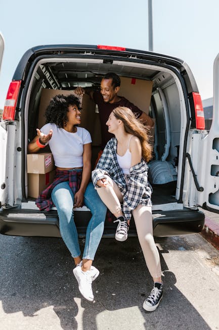 Two young women sit inside the open rear of a moving van parked on a paved street, engaging in conversation and smiling. One woman has curly black hair, wears a light blue T-shirt, and has her hand raised in a gesturing manner. The other has long, wavy brown hair, is dressed in a white top with a black-and-white checkered shirt tied around her waist, and is wearing black-and-white sneakers. Several cardboard boxes and packaging materials, such as plastic wrapping and moving blankets, are visible inside the van, indicating a home relocation. The van is part of a professional removals service, specifically from Man and Van Bow, providing furniture transport and packing and moving solutions. The environment is well-lit with clear weather, and the van is positioned near a curb, emphasizing an active loading or unloading process typical of house removal activities.