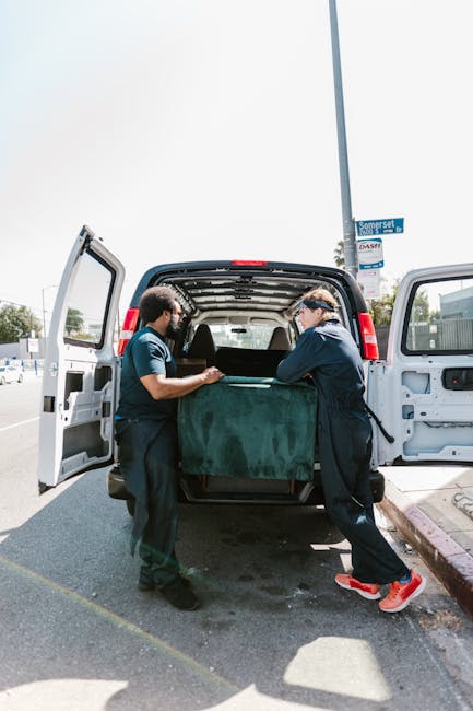 Inside a white moving van from Man and Van Bow, two movers are visible preparing for a home relocation. The driver, a man with a beard and short dark hair, is smiling at the camera, wearing a blue uniform with a logo on the chest, seated in the driver’s seat. Next to him, a woman with long dark hair, wearing a dark zip-up jacket, is seated in the passenger seat, also smiling. The van’s interior shows safety equipment such as seat belts, and the vehicle exterior displays the company's name, 'Moving Company,' along with the services offered, including local and long-distance moves. The scene is set outdoors with a background of urban buildings, and sunlight illuminates the scene, emphasizing the professional moving process involving packing supplies, such as cardboard boxes and protective blankets, inside the van, which is part of the furniture transport and packing and moving services provided by Man and Van Bow.