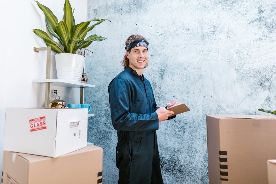 Close-up of a person in a dark t-shirt and jeans lifting a large cardboard box during a home relocation process, with a focus on their hands and torso. The box is sealed with packing tape and has a re