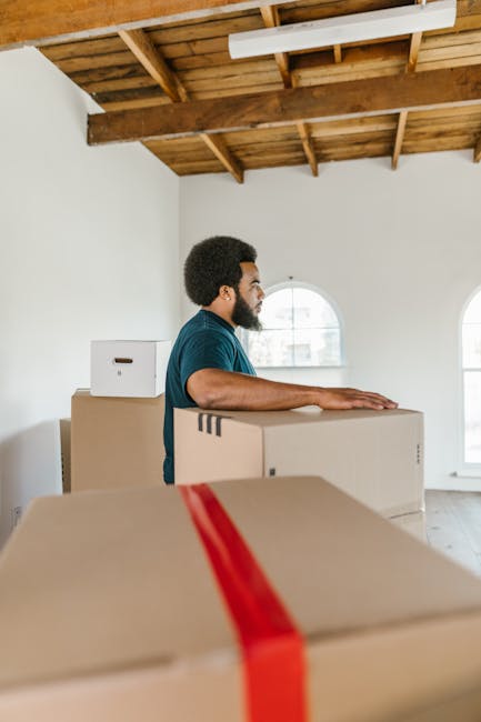 A person wearing a dark green T-shirt and dark trousers is carefully holding a large cardboard moving box with both hands. The box has a red and white caution label attached that reads 'CAUTION THIS S